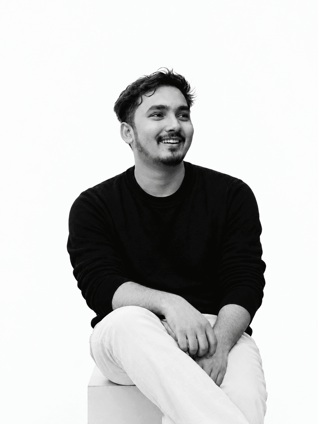 Sabbir Mohammad Sami, smiling and looking off to the side, seated on a white block in a black sweater — black-and-white studio portrait